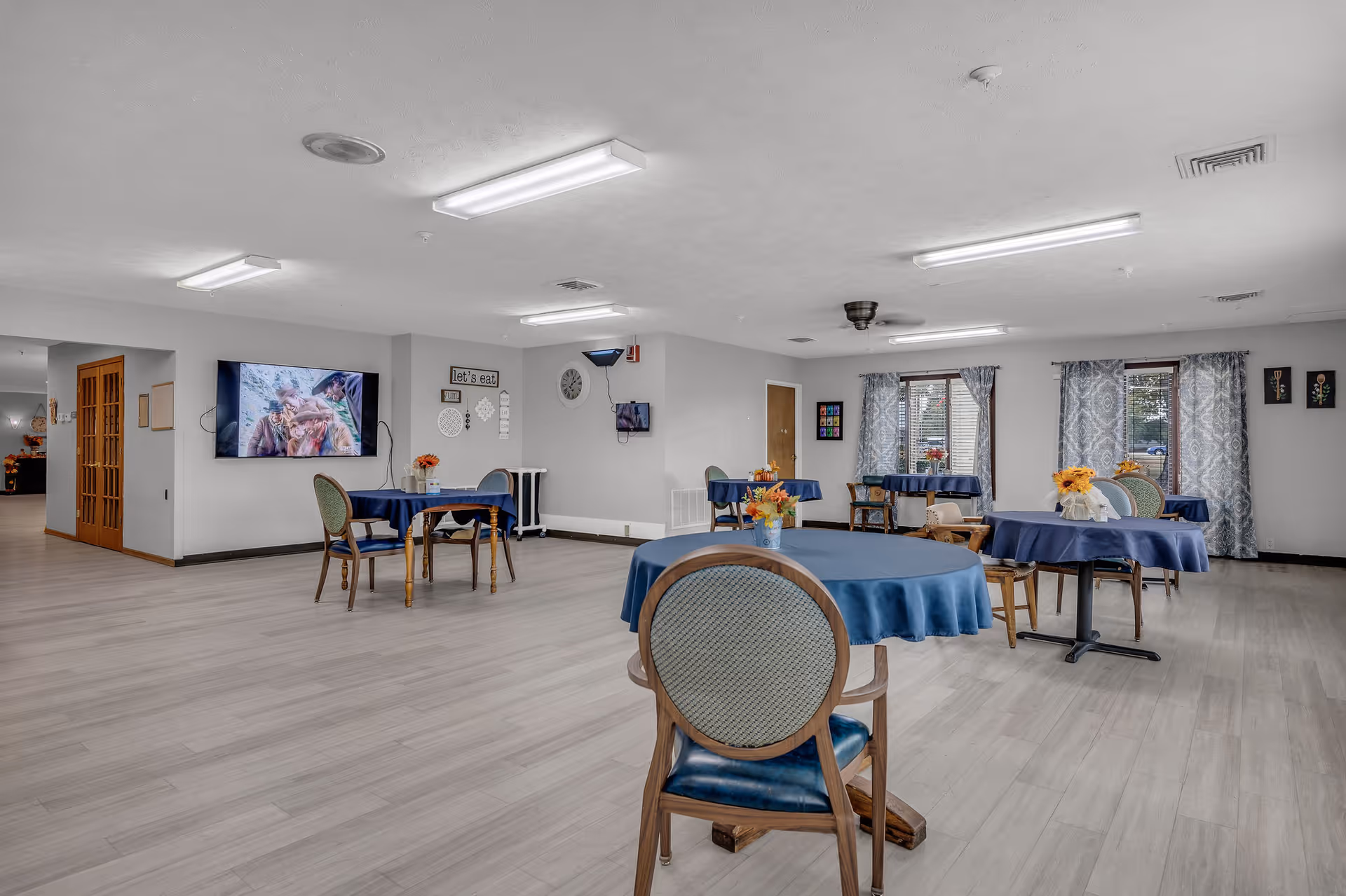 A spacious dining room with several round tables covered with blue tablecloths and decorated with small flower arrangements. The room has light-colored wooden flooring, large windows with patterned curtains, and fluorescent ceiling lights. A flat-screen TV is mounted on one wall, and there are a few chairs around the tables.