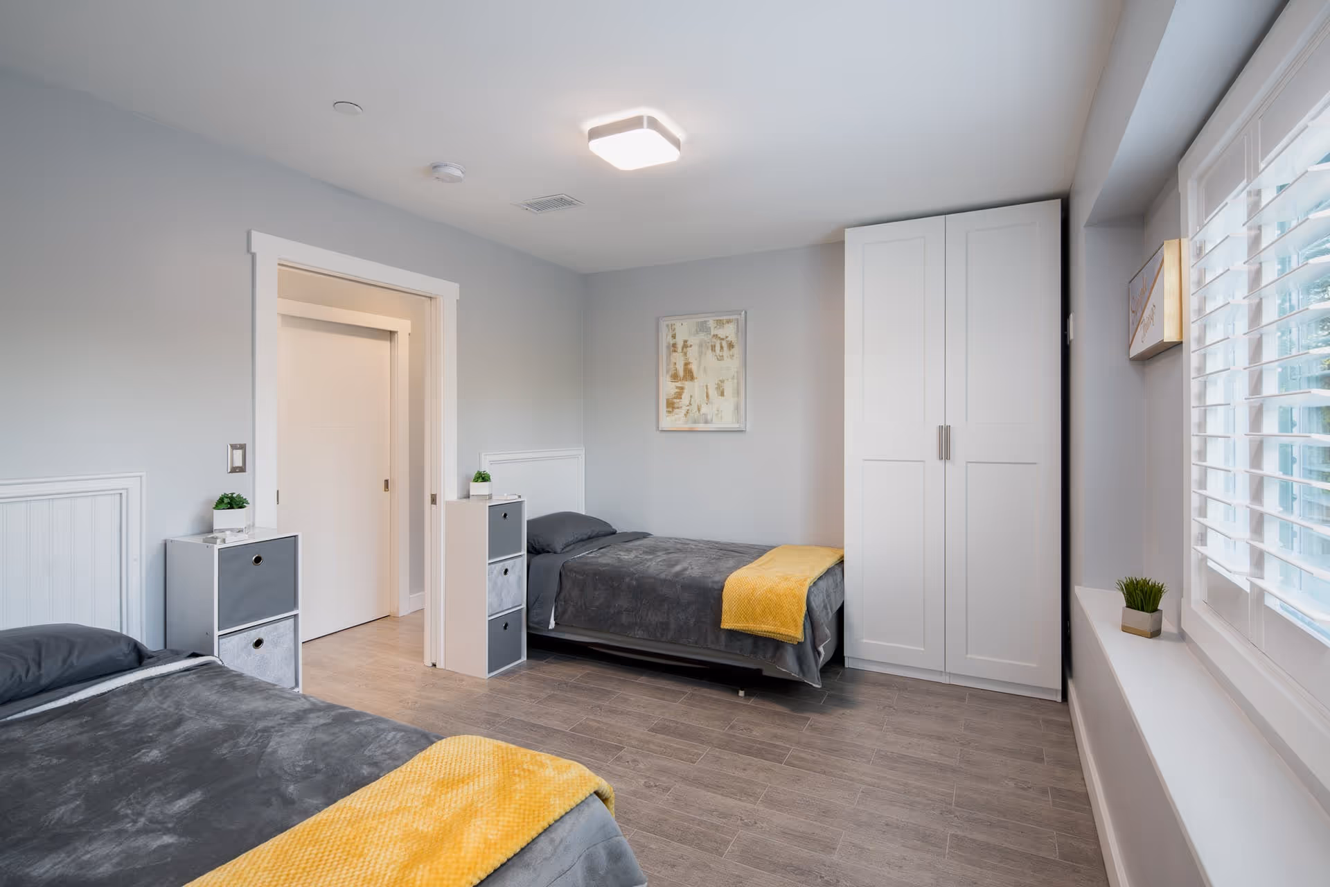 Bright, neatly arranged bedroom with two single beds in gray bedding and yellow throws, a white wardrobe, small storage units, and a window with shutters.