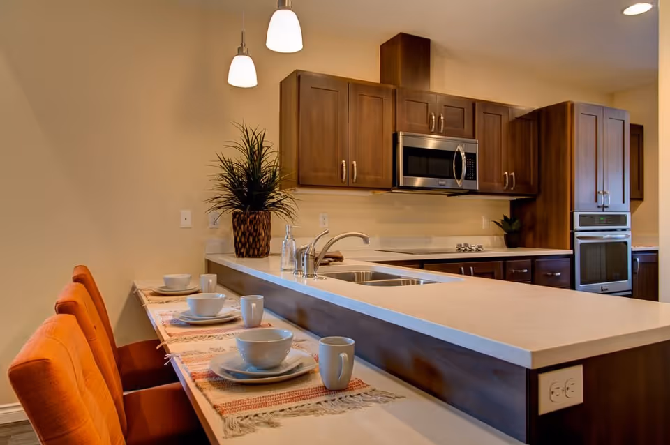Modern kitchen area with wooden cabinets, a built-in microwave, oven, and a stovetop. A countertop with a double sink and a breakfast bar with four orange cushioned chairs is set with bowls, plates, and mugs. Two pendant lights hang above the counter, and a potted plant is placed near the wall.