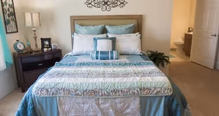 A neatly made bed with multiple pillows and a patterned quilt in a bedroom. There is a dark wooden nightstand with a lamp, clock, and picture frame on the left side of the bed. A door to a bathroom is open on the right side of the room, showing a toilet inside. The walls are light-colored, and there is a decorative metal wall art above the bed.