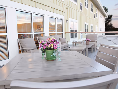 Outdoor patio deck with tables, chairs, potted flowers, and a light-colored building facade with windows.