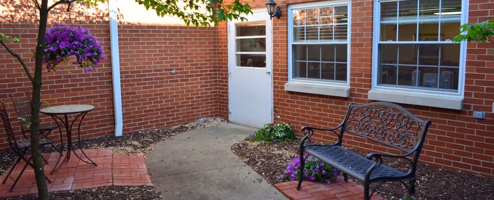 Outdoor courtyard area with a brick wall and two windows. There is a black metal bench with decorative backrest on the right side, a small round table with two chairs on the left, and a hanging basket of purple flowers. The ground has a concrete path and mulch with some small plants.