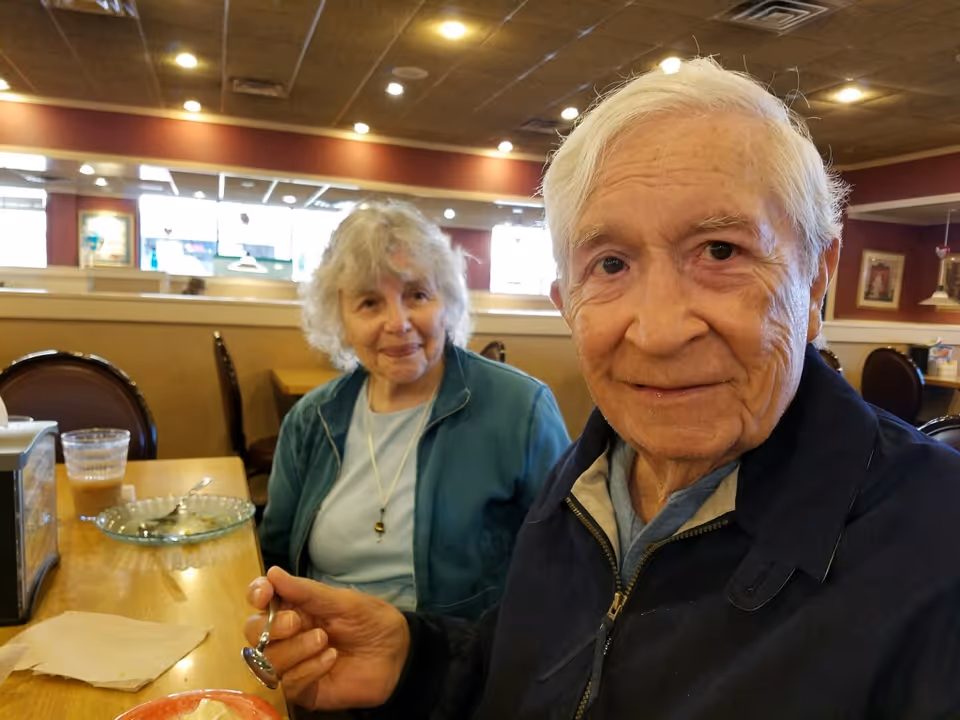 An elderly man and woman sitting at a dining table in a restaurant or dining area. The man is holding a spoon and looking at the camera, while the woman is smiling and looking at the man. There are plates and a drink on the table, and the background shows other tables and chairs with warm lighting.