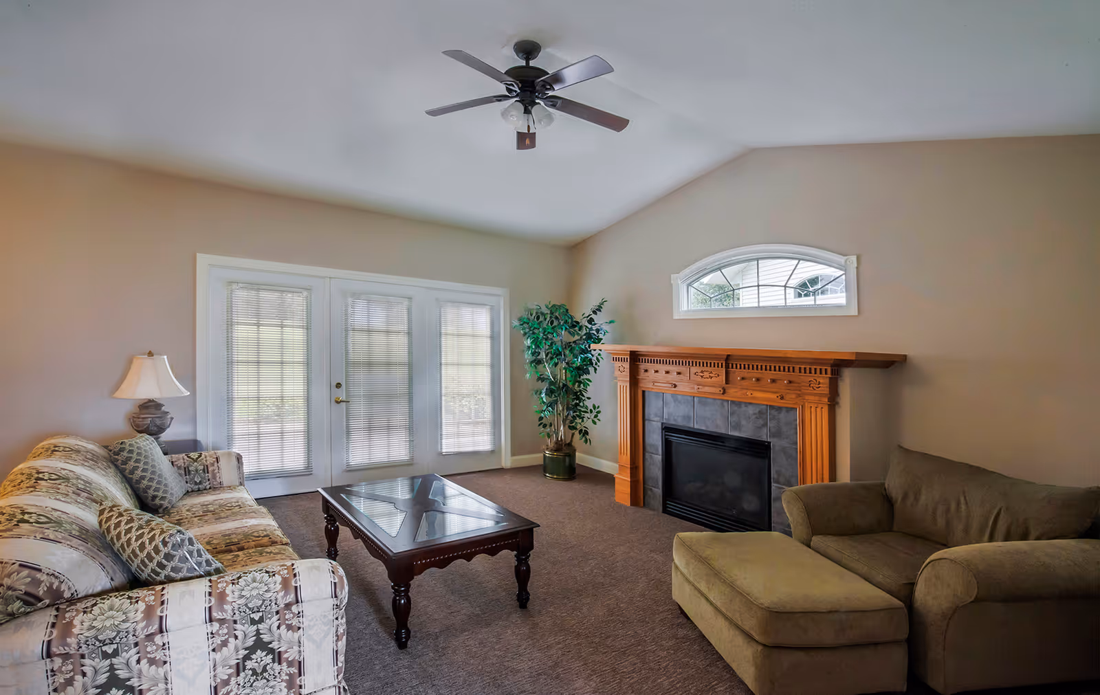 A cozy living room with a floral patterned sofa on the left, a wooden coffee table in the center, and a green armchair with an ottoman on the right. There is a wooden fireplace with a tiled surround on the far wall beneath a small arched window. A potted plant is placed next to the fireplace, and French doors with blinds lead outside. The ceiling has a ceiling fan with a light fixture.