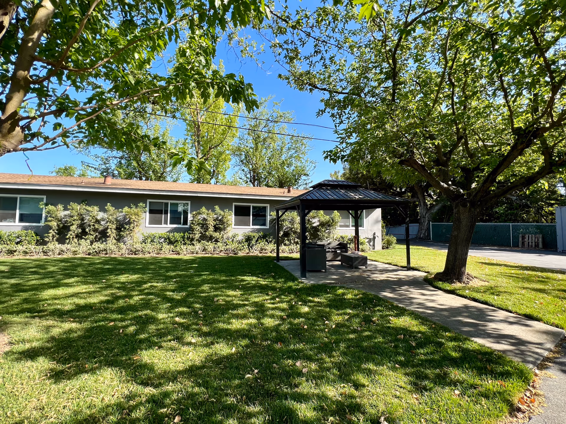 Grassy courtyard with a covered gazebo and seating in front of a single-story building surrounded by trees.