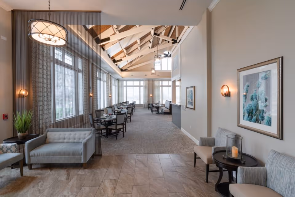 A spacious and well-lit dining area with large windows along one side, featuring multiple dining tables and chairs arranged neatly. The ceiling has exposed wooden beams and hanging light fixtures. There are comfortable seating areas with sofas and chairs near the entrance, along with framed artwork on the walls and a small table with a candle.