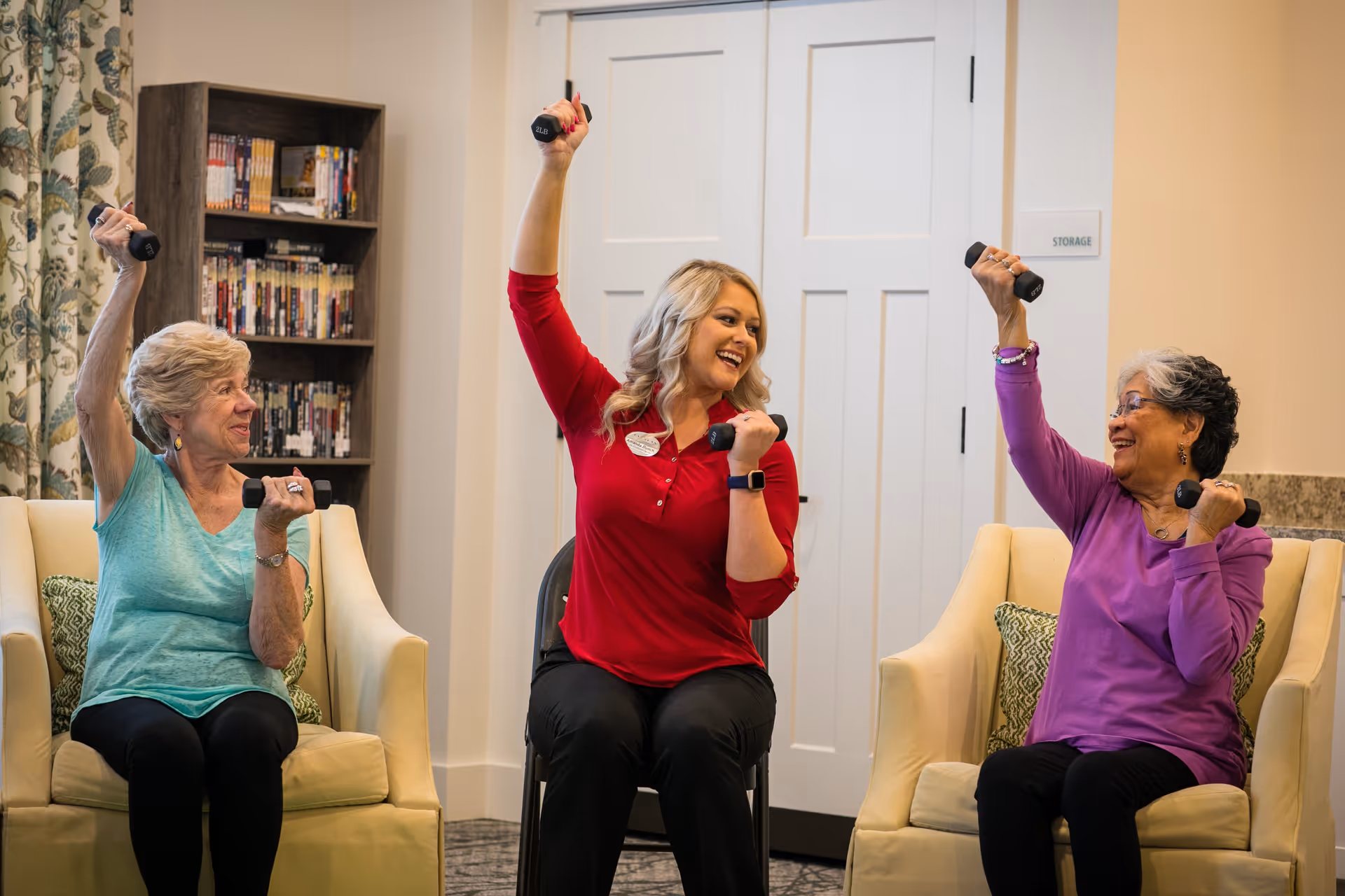 Two elderly women and a younger woman sitting in chairs indoors, smiling and lifting small dumbbells as part of a seated exercise activity. The room has a bookshelf with DVDs and a door labeled 'Storage' in the background.