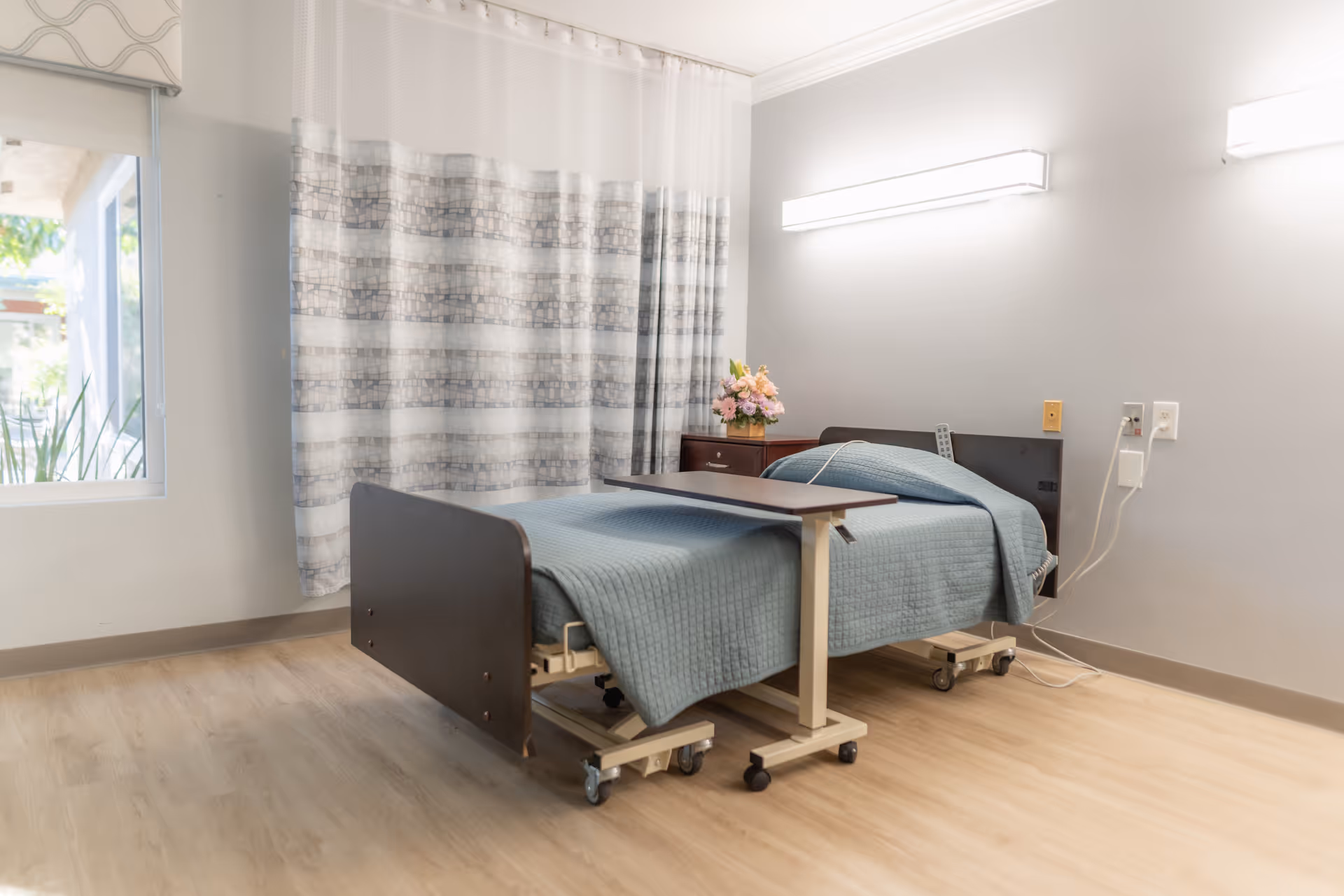 A clean hospital-style single bed with blue bedding in a bright senior living resident room with a bedside table, flowers, and a window.
