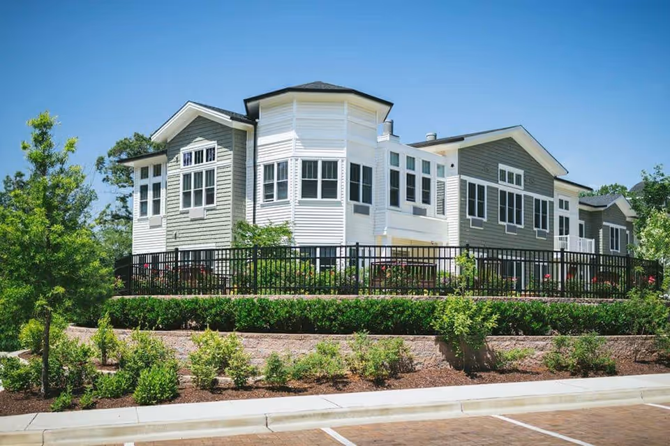 Exterior view of a modern senior living facility building with white and gray siding, multiple windows, surrounded by landscaped greenery and a black metal fence under a clear blue sky.