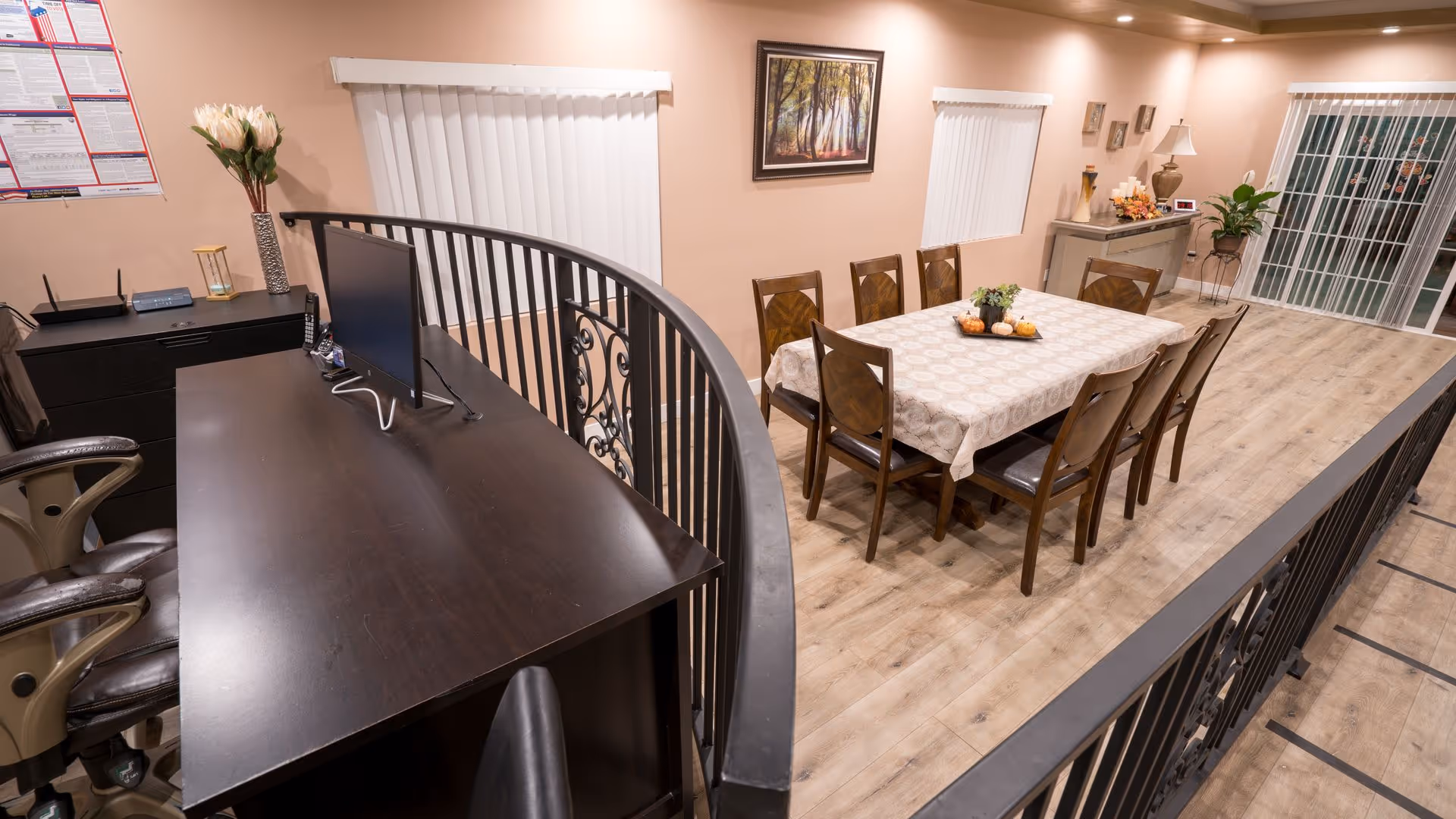 Interior view of a senior living dining area with a long table and chairs and a nearby desk with a computer behind a decorative railing.