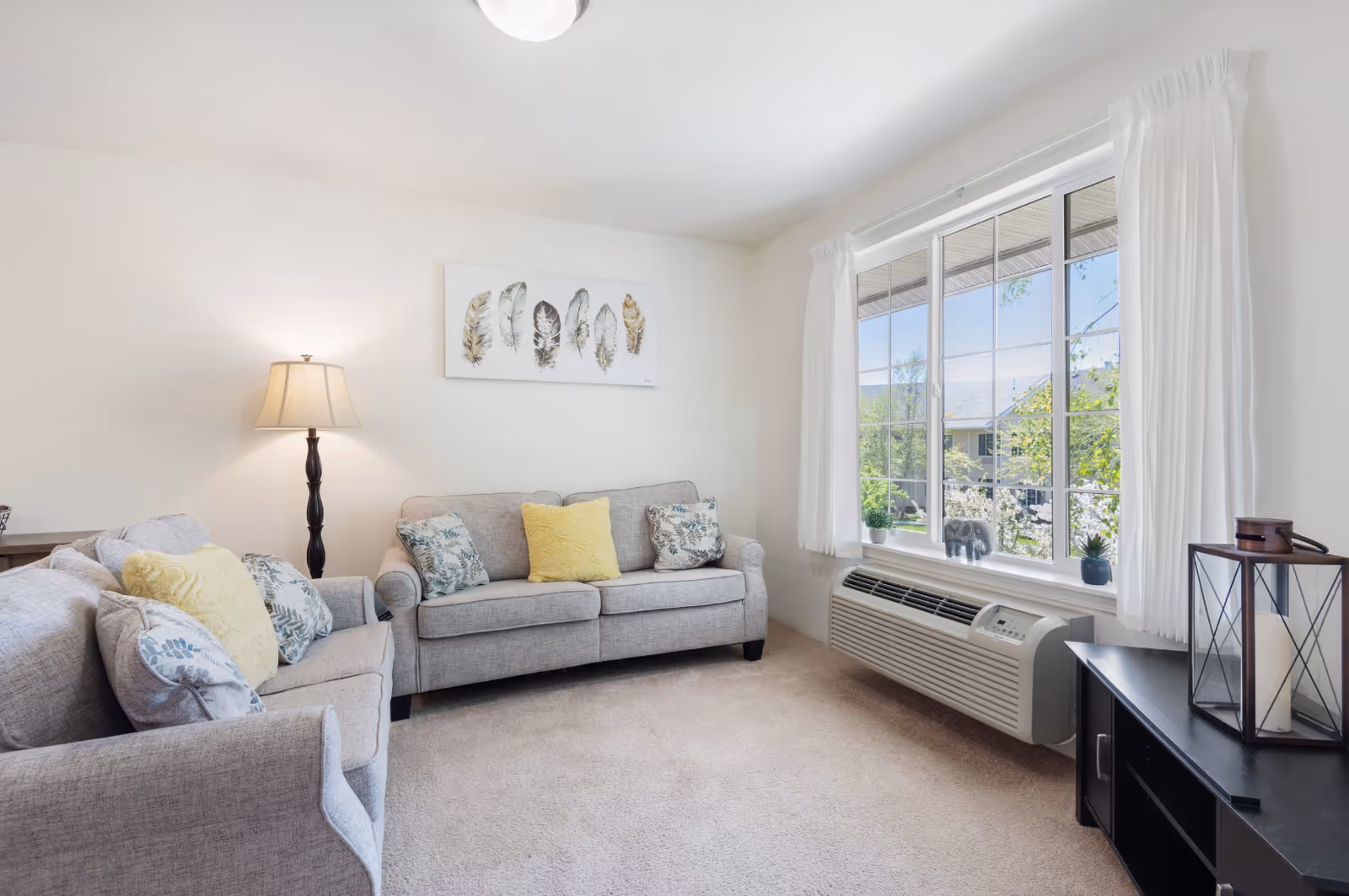 A bright and cozy living room with two gray sofas adorned with patterned and yellow pillows, a floor lamp, a large window with white curtains letting in natural light, a black TV stand with a decorative lantern, and a wall art featuring feathers.