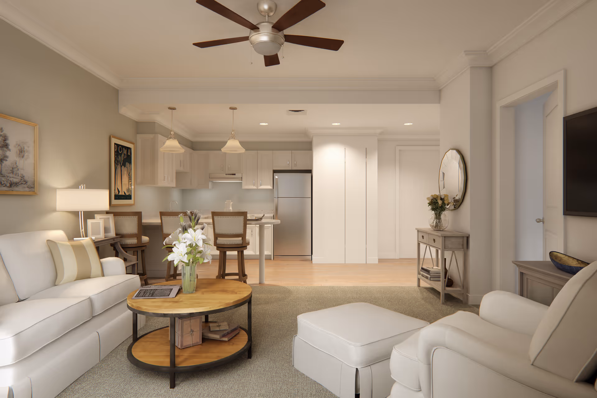 A bright and cozy living room area with a white sofa, armchair with ottoman, and a round wooden coffee table with a vase of white flowers. The room opens into a kitchen with white cabinets, a stainless steel refrigerator, and a small dining area with three chairs. The walls are painted light beige, and there is a ceiling fan above. Decorative elements include framed artwork, a table lamp, and a round mirror above a small console table with flowers.