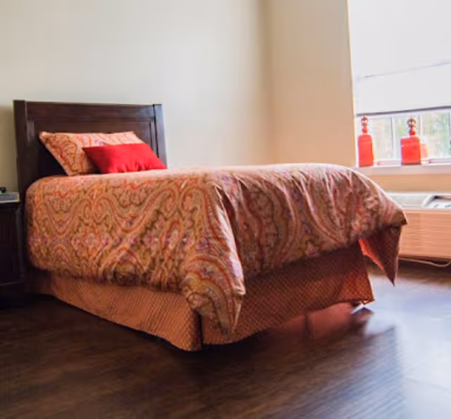 A bedroom with a single bed featuring a patterned bedspread and two pillows, one red and one matching the bedspread. The room has wooden flooring, a window with white blinds, and two red decorative items on the windowsill.