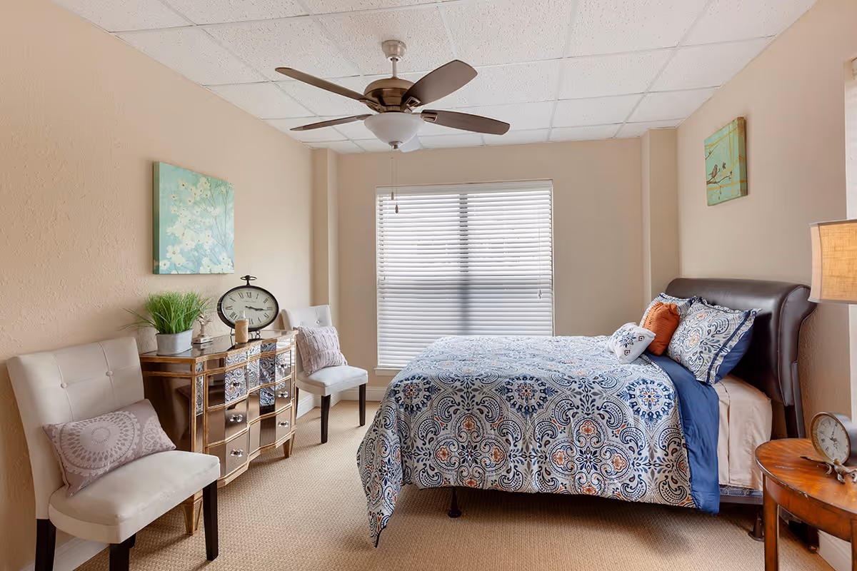 A cozy bedroom with a patterned blue and white bedspread on a dark wooden bed frame. The room features beige walls, a ceiling fan with light, a window with white blinds, two white chairs with decorative pillows, a mirrored dresser with a clock and plant on top, and two pieces of wall art.