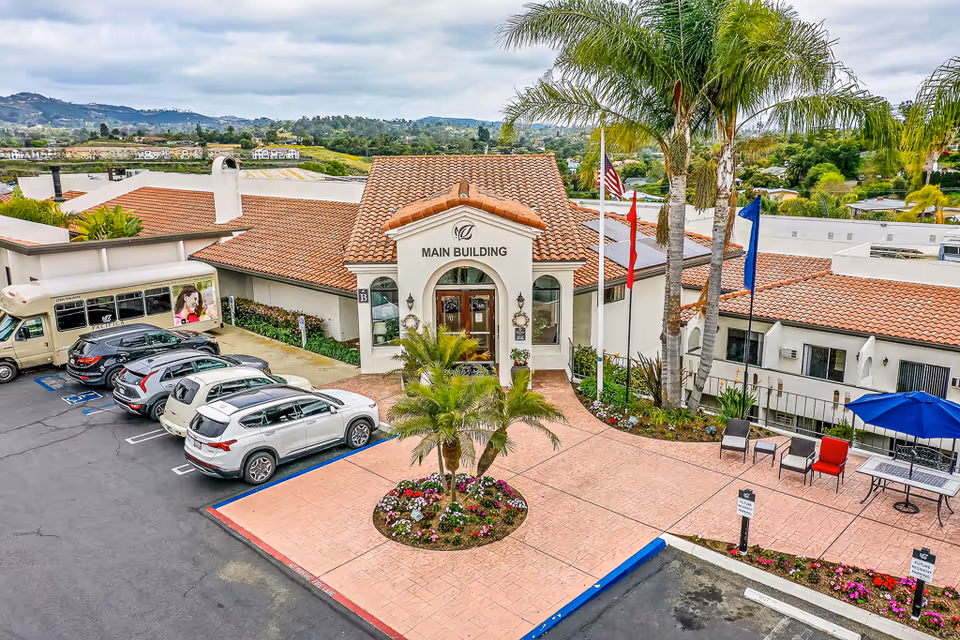 Front entrance of Rancho Vista Senior Living main building with a terracotta roof, flagpoles, palm trees, parked cars and a paved courtyard.