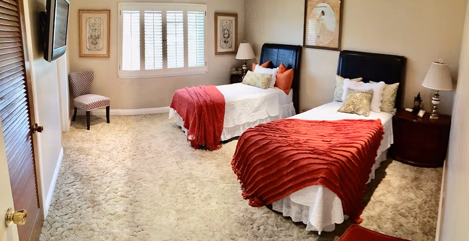 A bedroom with two twin beds each covered with white bedding and red throws. Each bed has multiple pillows and a black headboard. There is a wooden nightstand with a lamp between the beds and another lamp on a small table on the right side. A patterned chair is placed near a window with white shutters. The room has beige walls and carpeted floor.