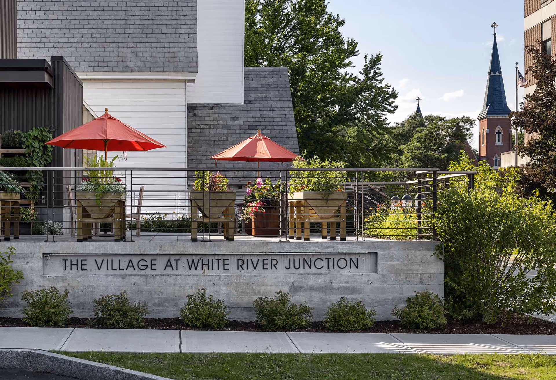 Outdoor patio area at The Village at White River Junction with red umbrellas, wooden planters filled with plants and flowers, surrounded by greenery and buildings in the background including a church steeple.