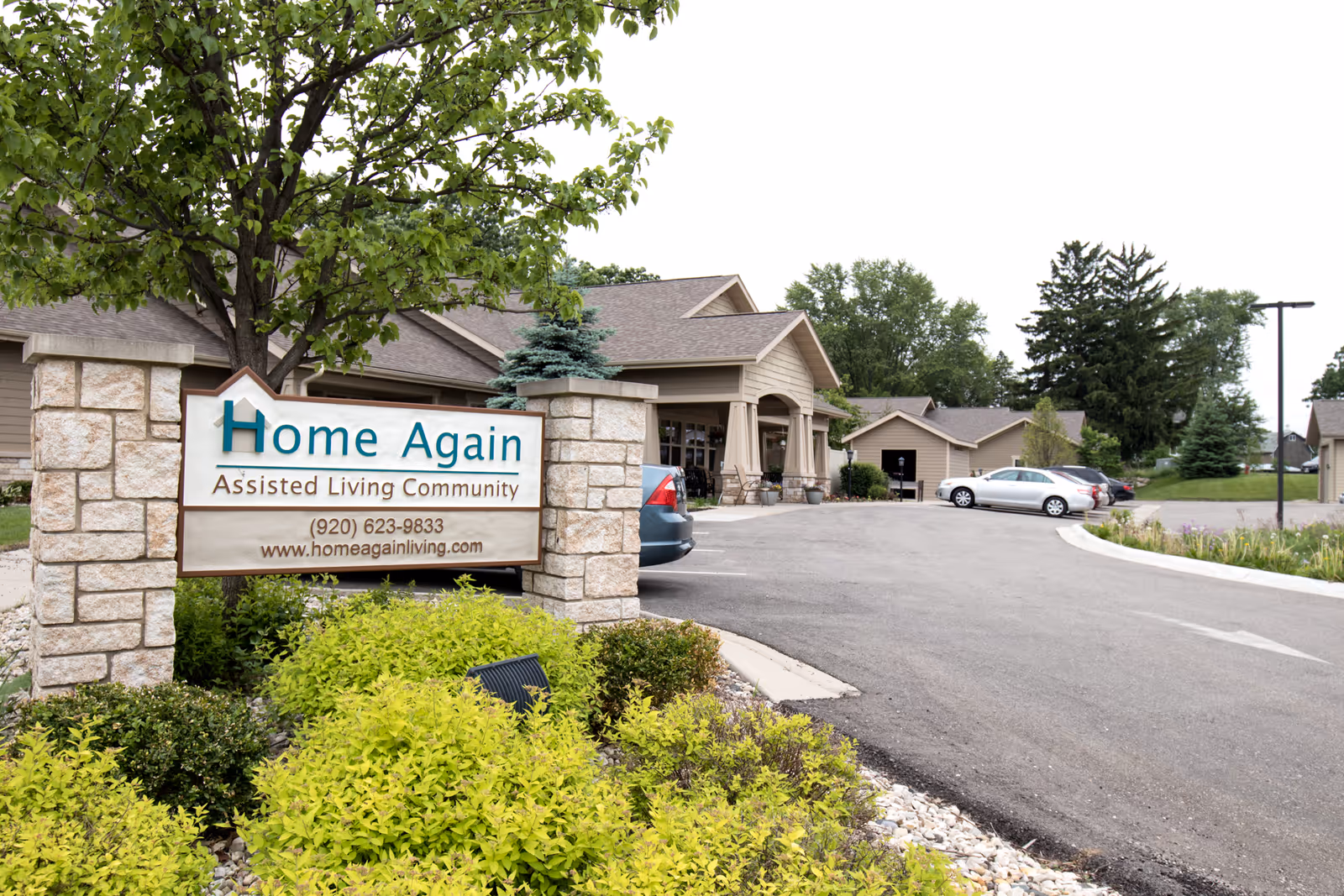 Exterior view of Home Again Assisted Living Community showing the entrance driveway, parked cars, and a sign with the facility's name, phone number, and website. The building is surrounded by greenery and trees.