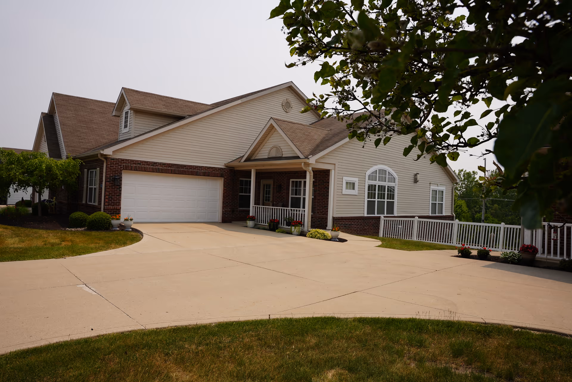 Exterior view of a single-story assisted living facility building with beige siding and brick accents, a large driveway, a white garage door, a small covered porch with potted plants, and a white fence. Trees and greenery surround the building.