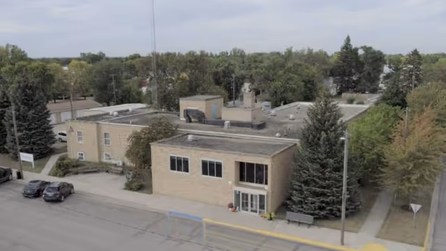 Front exterior view of a low-rise brick senior living community building with entrance, benches, parked cars, and surrounding trees.