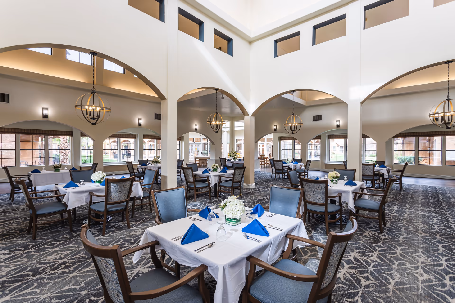 Spacious dining room with tables set with white linens and blue napkins under high arched ceilings and chandeliers.