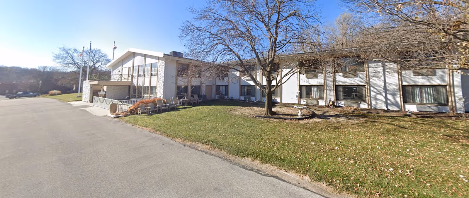Exterior view of a two-story building with large windows and a sloped roof, surrounded by a grassy area with a leafless tree in front. There is a paved driveway or parking area to the left, and several flagpoles near the entrance of the building under a clear blue sky.