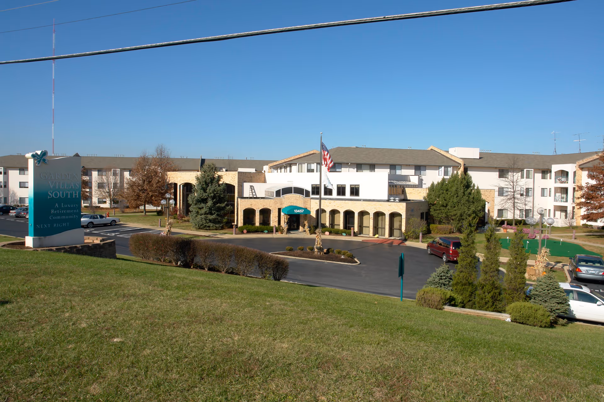 Exterior view of Garden Villas South, a luxury retirement community, showing a large multi-story building with a parking lot, landscaped greenery, and an American flag in front under a clear blue sky.