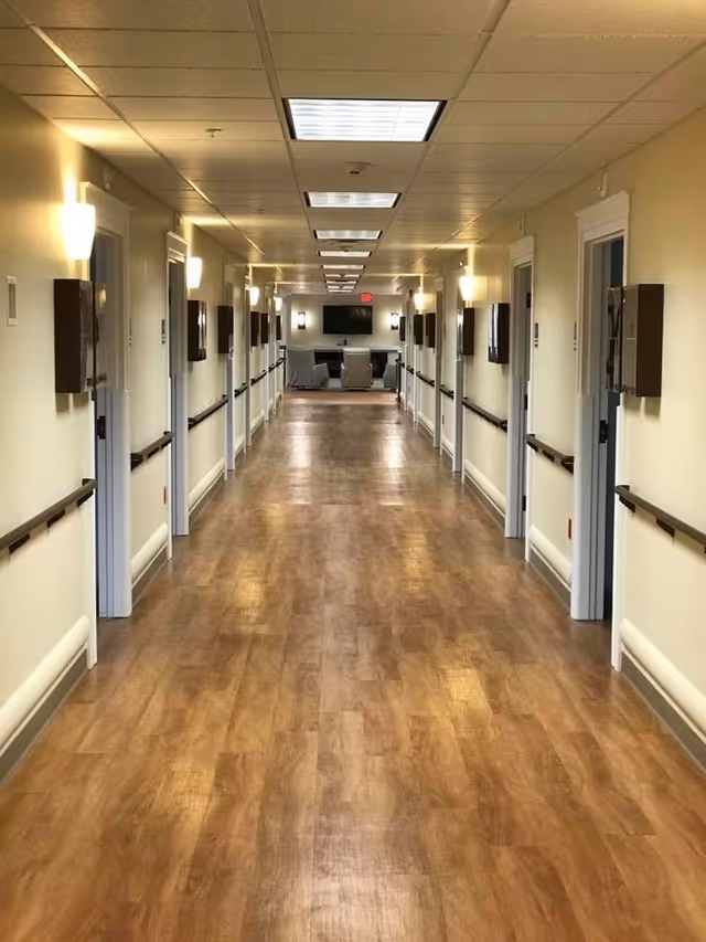 Long hallway in a senior living facility with wooden flooring, beige walls, handrails on both sides, wall-mounted lights, and multiple doorways leading to rooms. At the end of the hallway, there is a seating area with chairs and a wall-mounted television.