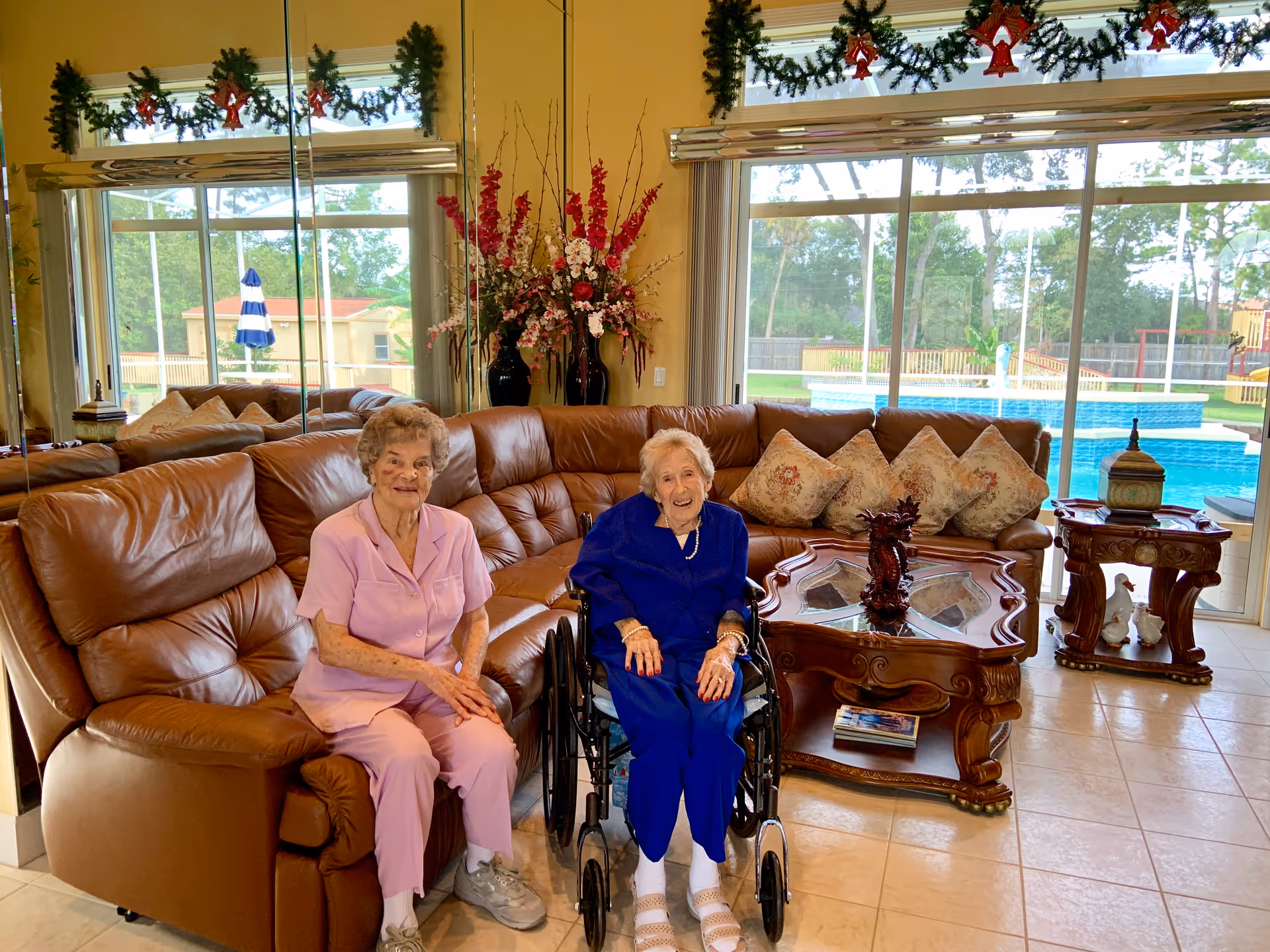 Two elderly women sitting in a living room with a large brown leather sectional sofa. One woman is seated on the sofa wearing a pink outfit, and the other is in a wheelchair wearing a blue outfit. Behind them is a glass sliding door showing a swimming pool and outdoor area. The room is decorated with floral arrangements, garlands with red bows above the windows, and a wooden coffee table with decorative items.