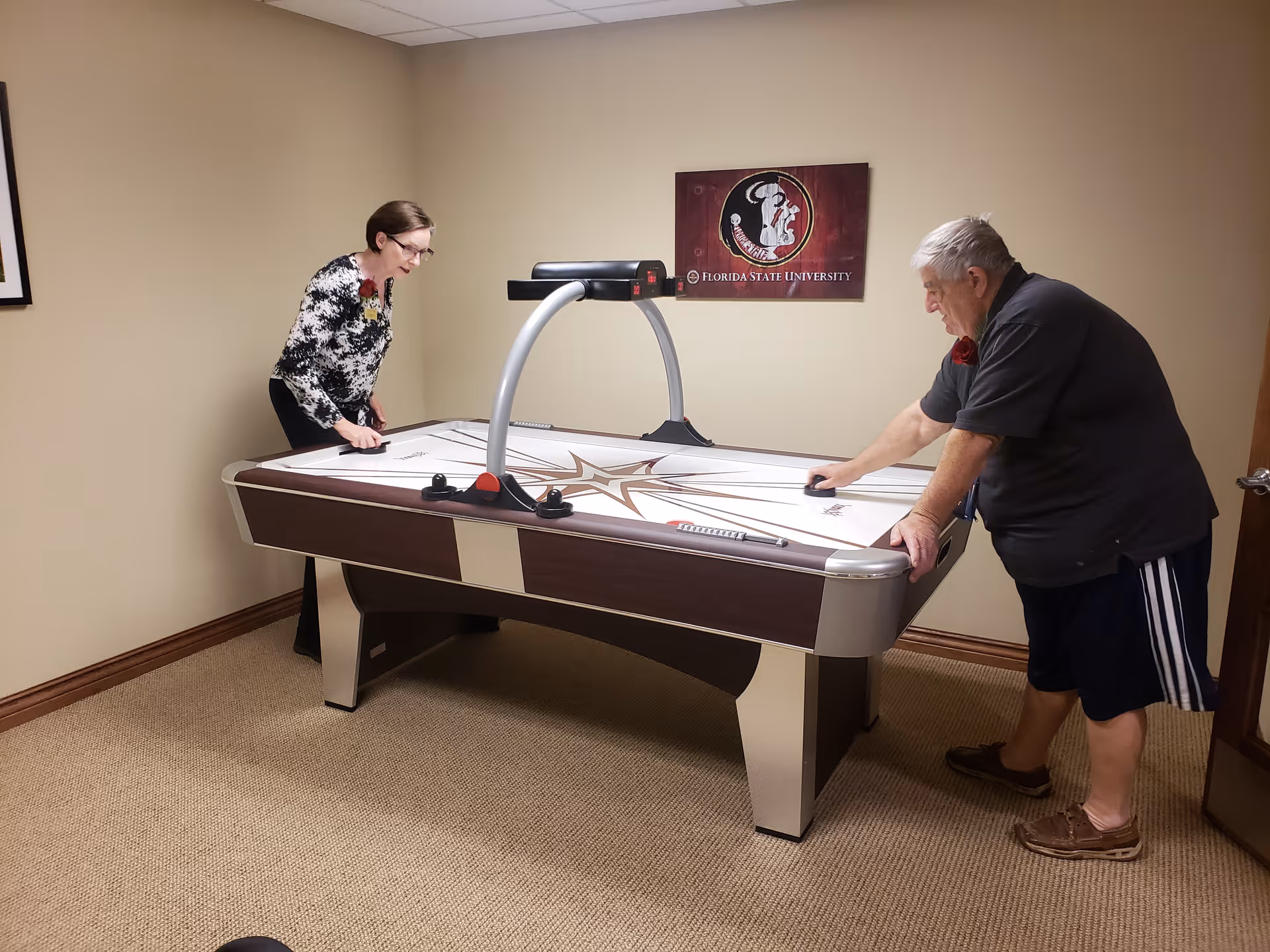 Two elderly people playing air hockey in a small room with beige walls and carpet. A Florida State University sign is mounted on the wall behind the air hockey table.
