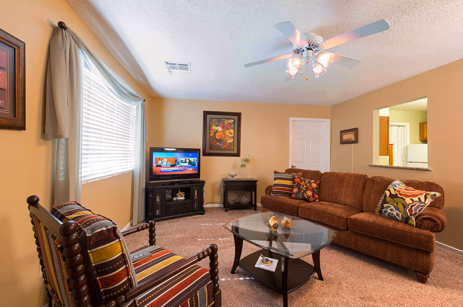 A cozy living room with a brown sofa adorned with colorful patterned pillows, a glass coffee table with decorative birds, a striped cushioned wooden chair, a TV on a black stand, a framed floral painting on the wall, beige walls, a window with light curtains, and a ceiling fan with lights.