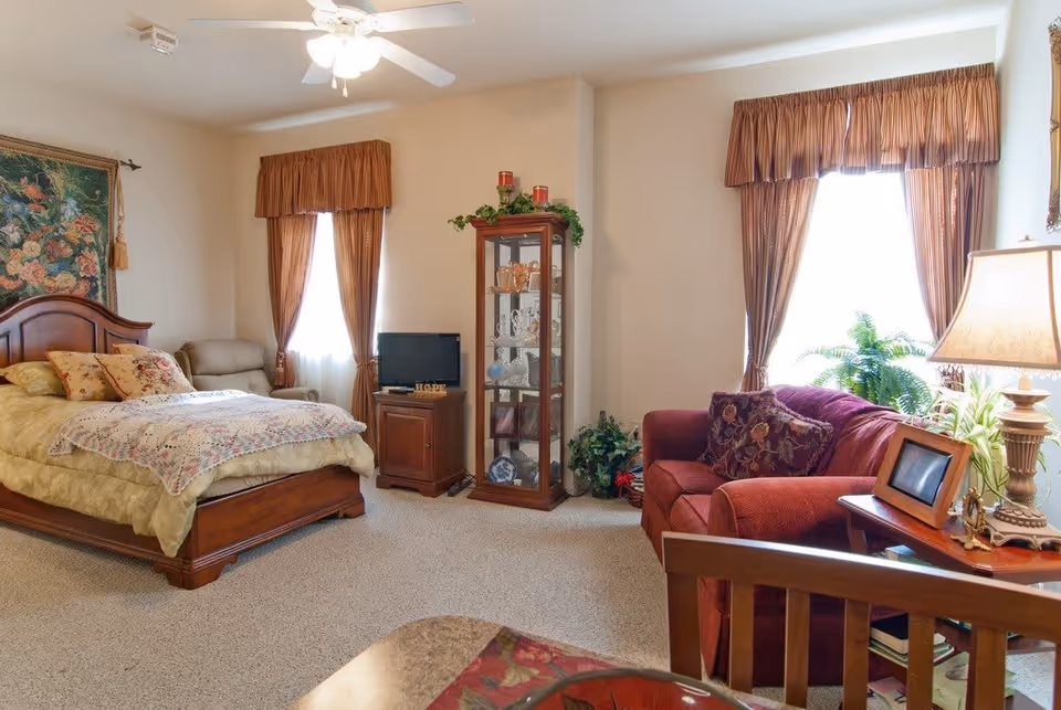 A cozy bedroom with a wooden bed featuring a floral quilt and pillows, a beige armchair near a window with brown curtains, a wooden cabinet with a small TV on top, a glass display cabinet with decorative items, and a red upholstered sofa with patterned cushions next to a side table with a lamp and framed photo. The room has beige walls and carpeted floor.