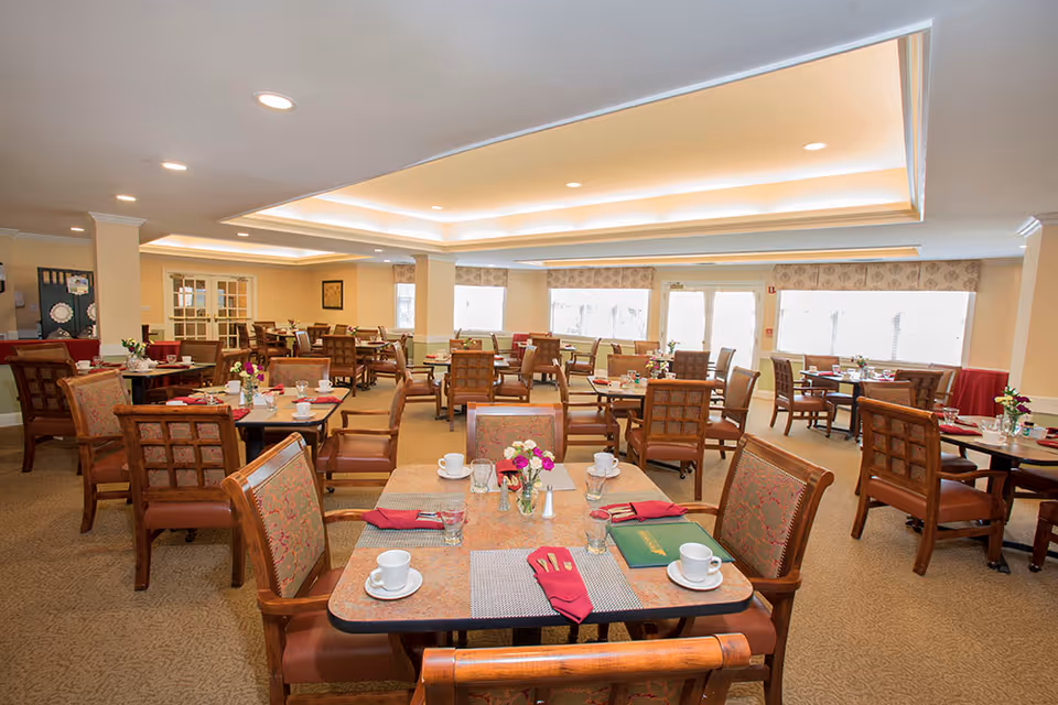 Bright, spacious dining room with multiple set tables and upholstered chairs in a senior living facility.