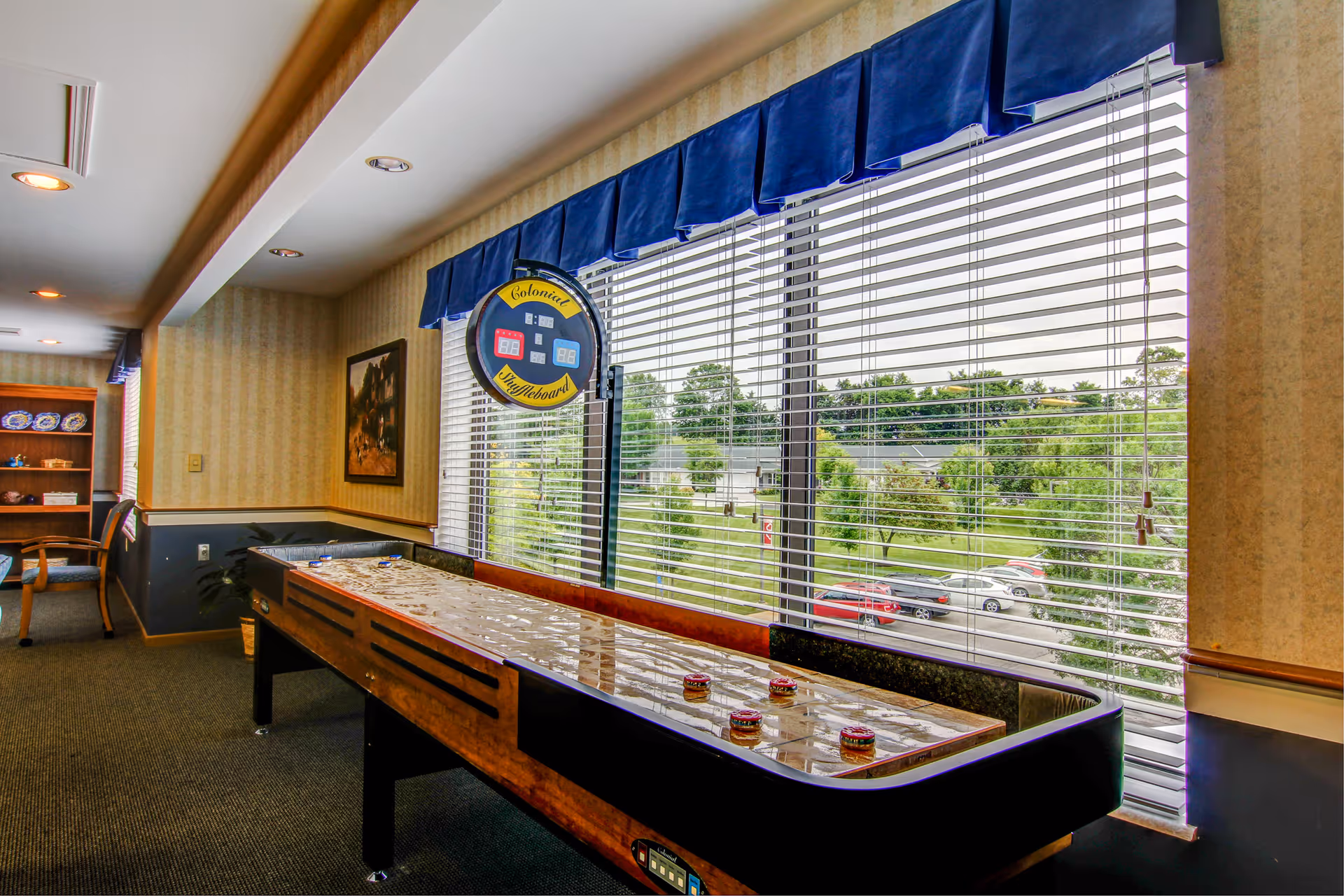 A shuffleboard table in a well-lit room with large windows covered by blinds and blue valances. Outside the windows, there is a view of trees, grass, and parked cars. The room has carpeted floors, wallpapered walls, and a framed picture on the wall. There is also a wooden shelving unit with decorative plates and chairs in the background.