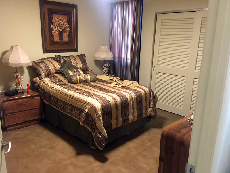 A bedroom with a bed covered in a striped and floral patterned comforter and multiple pillows. There are two bedside tables, each with a decorative lamp. A framed floral painting hangs on the wall above the bed. A window with striped curtains is visible next to a closet with sliding louvered doors.