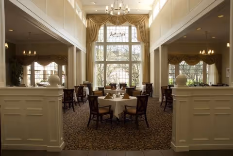 Elegant dining room with tables and chairs set for meals, chandeliers, and large windows with draped curtains.