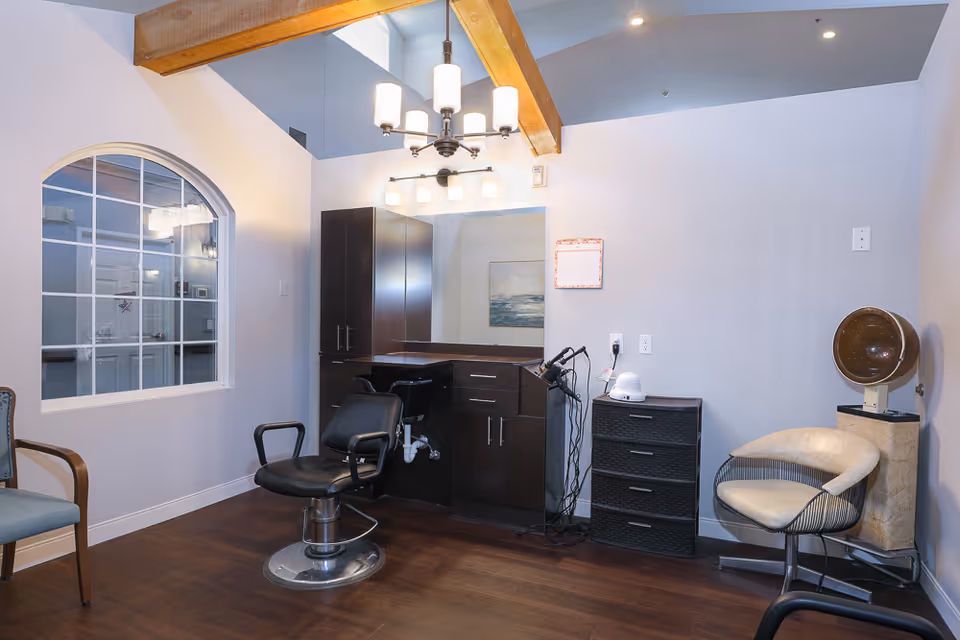 Interior view of a hair salon area in an assisted living facility featuring a black salon chair in front of a large mirror with dark wood cabinetry. There is a chandelier and wall light fixtures above, a window with white framing on the left wall, and a vintage hair dryer on a stand next to a cream-colored chair on the right side.