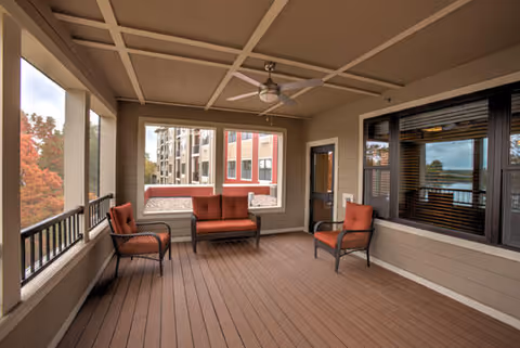 A covered outdoor patio area with wooden flooring and beige walls. The space is furnished with a red cushioned loveseat and two matching armchairs. Large windows and open railings provide views of nearby buildings and trees with autumn foliage. A ceiling fan is mounted on the ceiling.