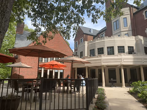 Outdoor patio area with tables and red umbrellas beside a multi-story brick and stucco senior living building.