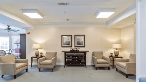 A cozy sitting area with four beige armchairs arranged around two small wooden side tables with lamps and plants. In the center against the wall is a dark wooden cabinet with decorative items and two framed pictures hanging above it. The room has soft lighting and a neutral color palette.