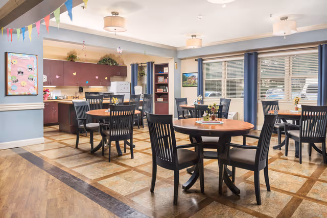 Communal dining room with round wooden tables, black chairs, large windows, and a kitchenette in the back.