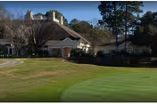 View of a senior living facility building named The Cypress of Hilton Head, surrounded by trees and greenery with a well-maintained lawn and putting green in the foreground.