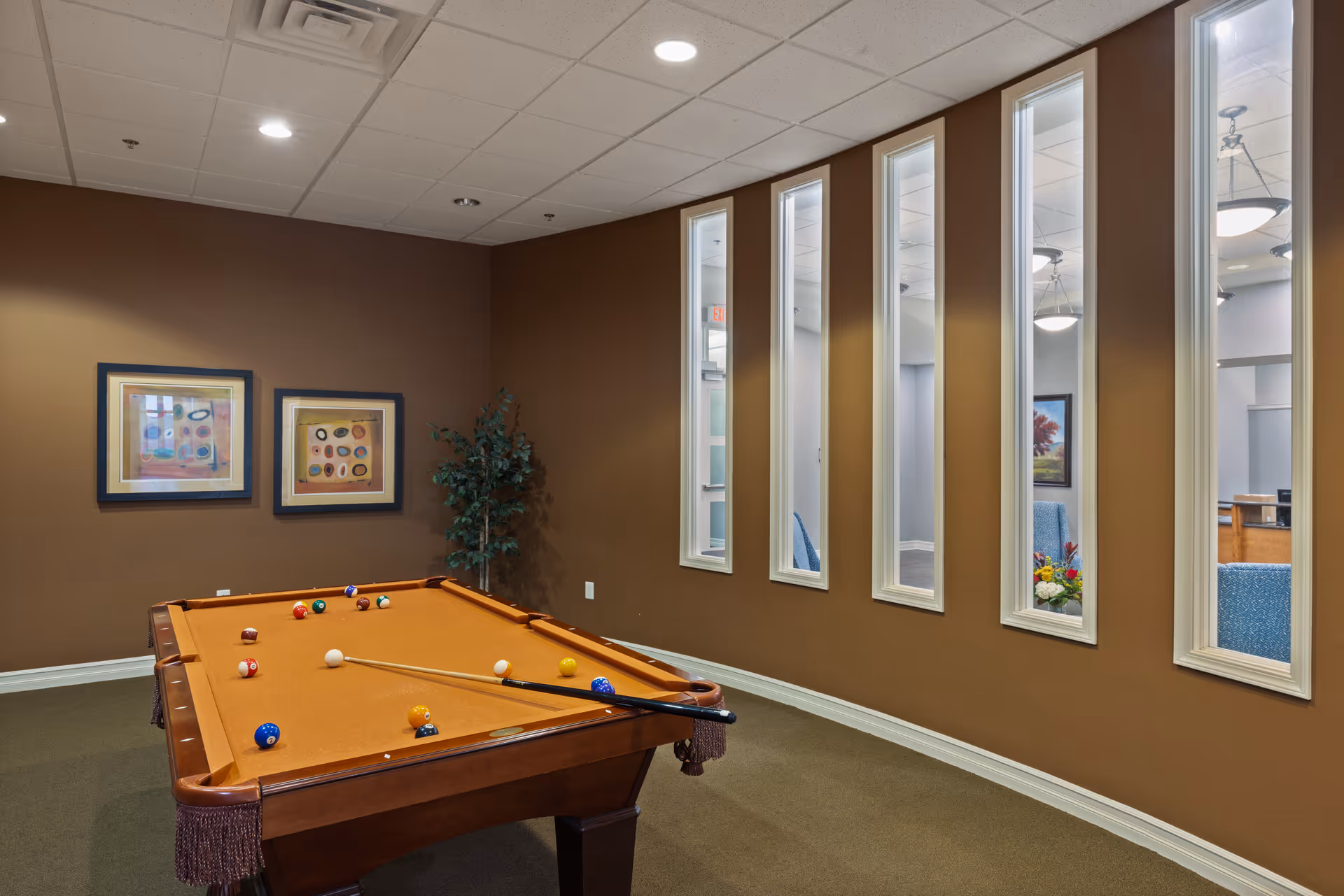 Interior room with a brown pool table with balls and cues set up, brown walls with two framed abstract paintings, a potted plant in the corner, and a series of tall narrow windows looking into an adjacent room with blue chairs and a floral arrangement.