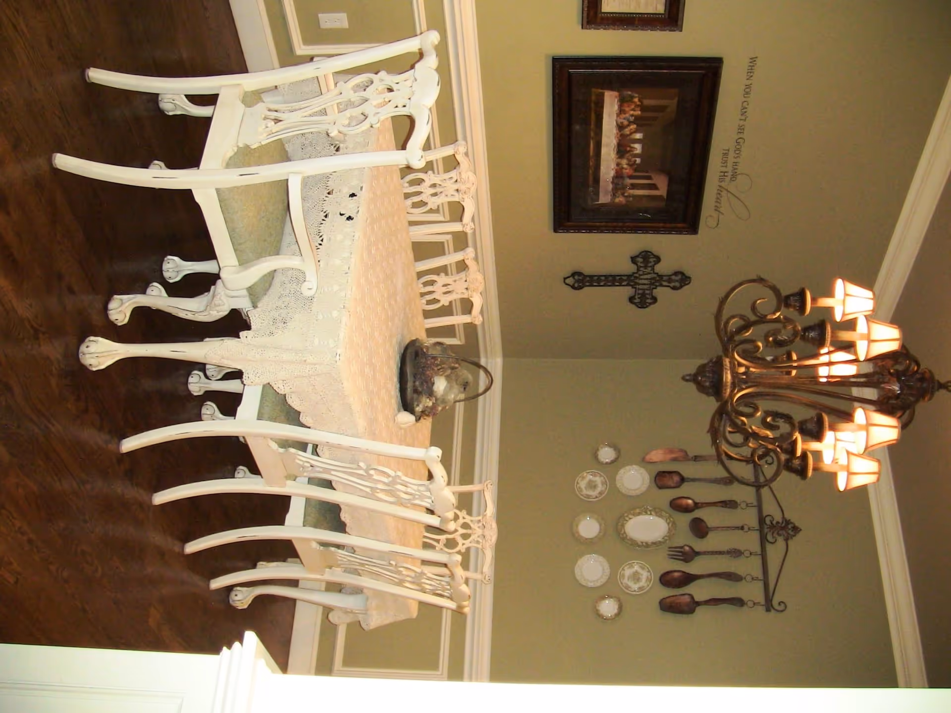 A dining room with a round table covered by a lace tablecloth and surrounded by six white ornate chairs. The floor is wooden, and the walls are painted light green with white trim. On the wall, there is a decorative cross, a framed picture, and a collection of decorative plates and large wooden utensils. A chandelier with multiple lights hangs above the table.