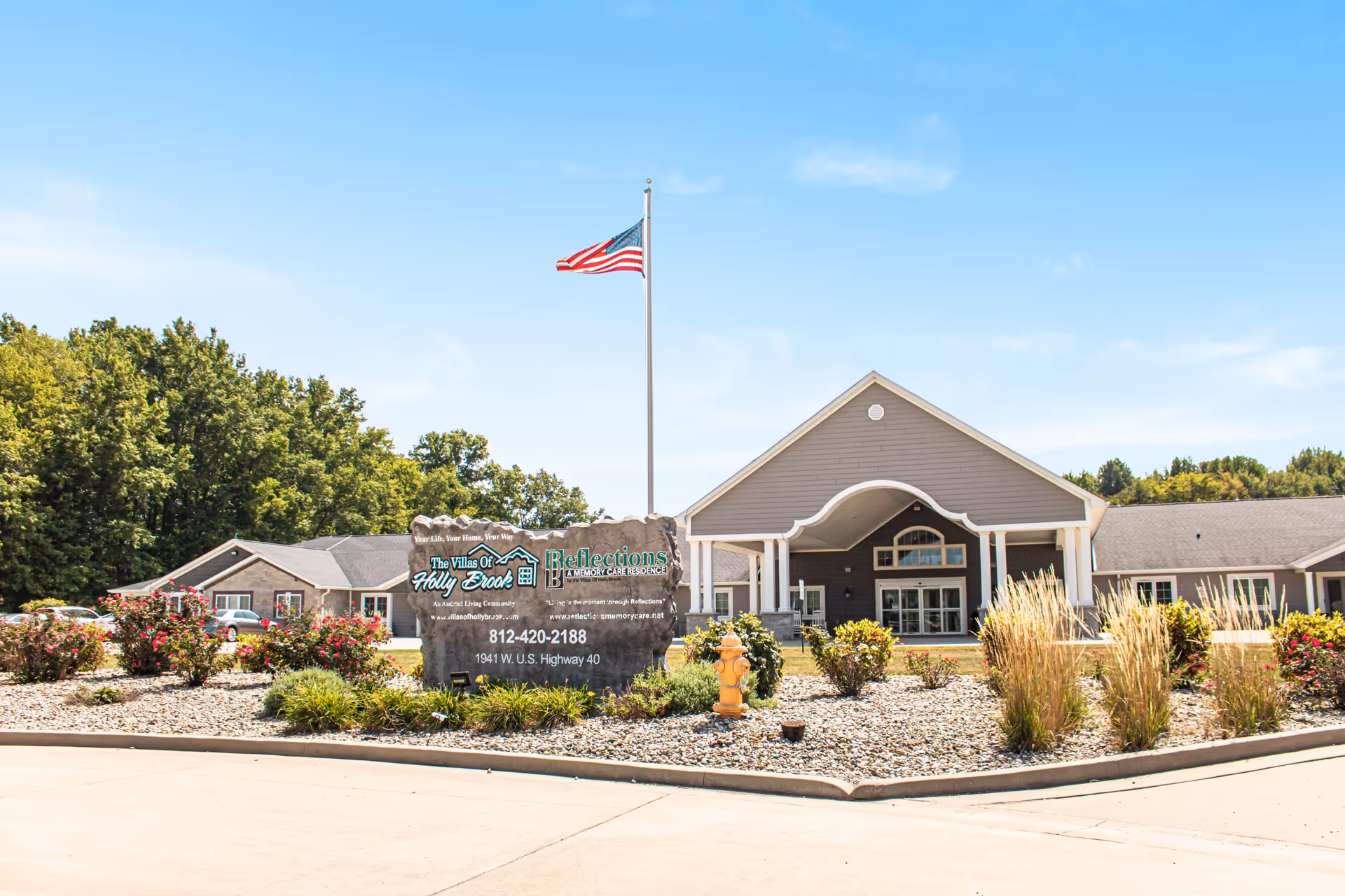 Exterior view of Villas of Holly Brook Assisted Living & Memory Care facility in Brazil, Indiana, showing a large building with a peaked roof, an American flag on a flagpole, landscaped garden with bushes and flowers, and a stone sign with the facility's name and contact information.