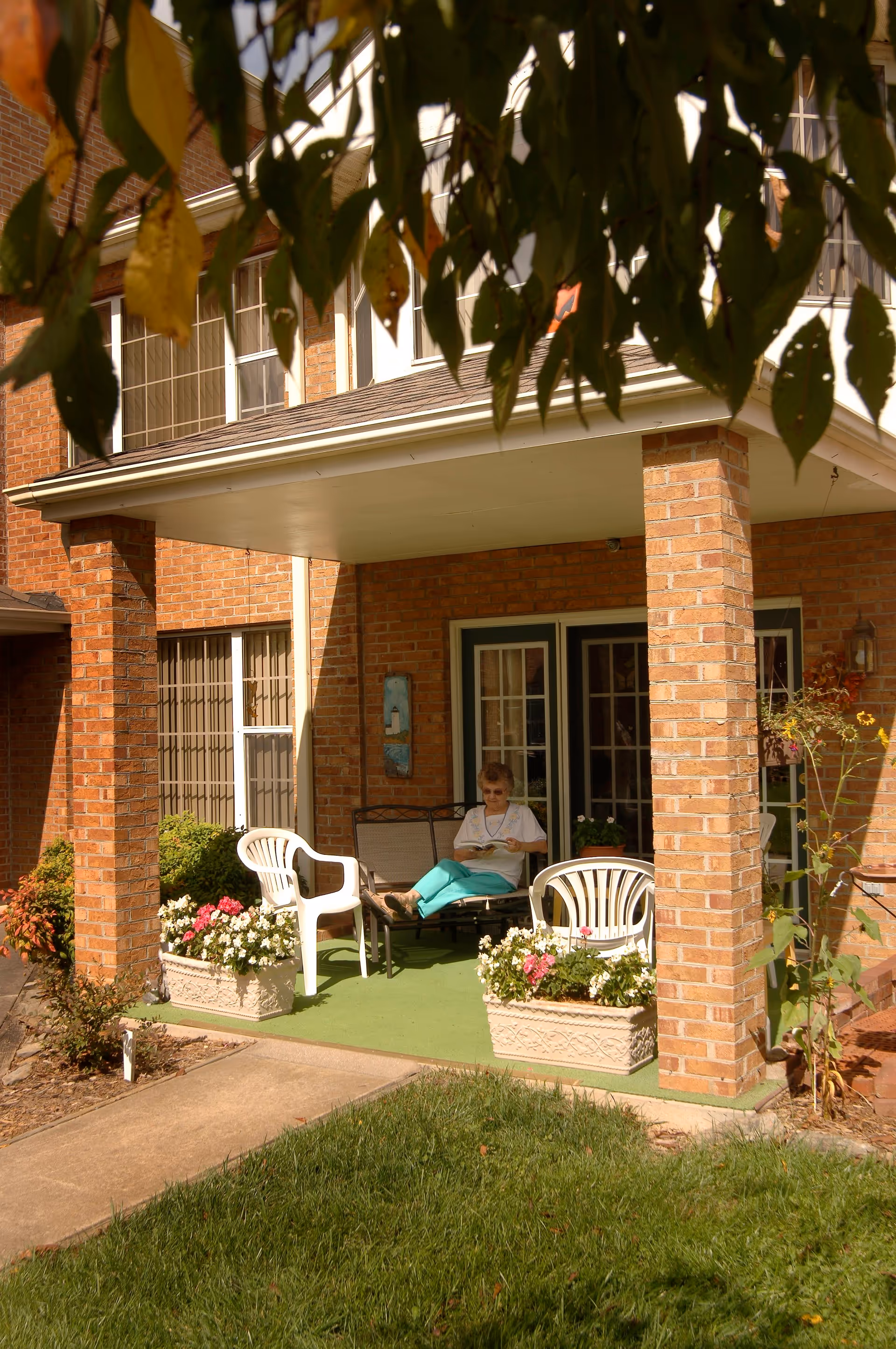 A covered brick porch with a person seated reading among white plastic chairs and flower planters.