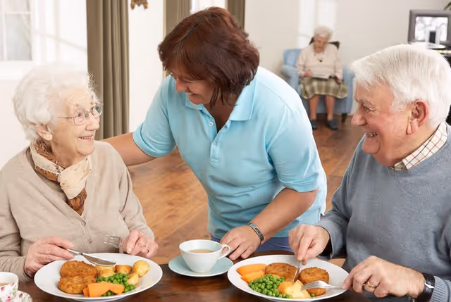 A caregiver in a light blue shirt smiling and interacting with an elderly woman and an elderly man who are seated at a table with plates of food including peas, carrots, potatoes, and breaded items. In the background, another elderly woman is seated in a chair reading a book in a well-lit room with wooden flooring.