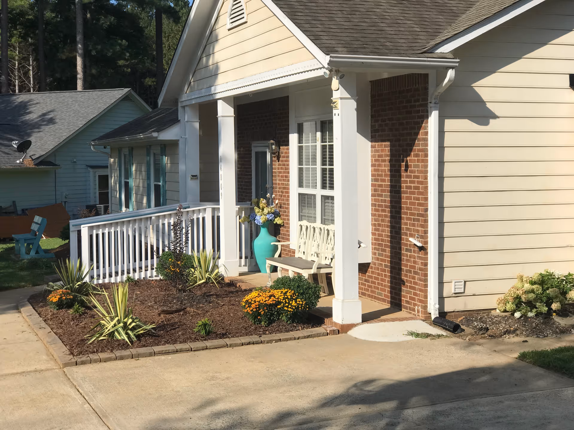 Front exterior view of a senior living facility building with a small porch, white railing, a bench, a large turquoise vase with flowers, and a landscaped garden bed with various plants and flowers.