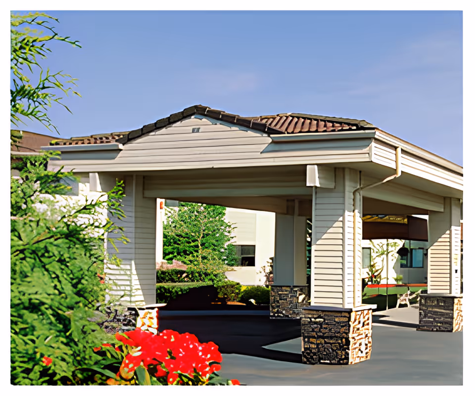 Covered entrance area of a building with white siding and stone pillars, surrounded by green bushes and red flowers under a clear blue sky.