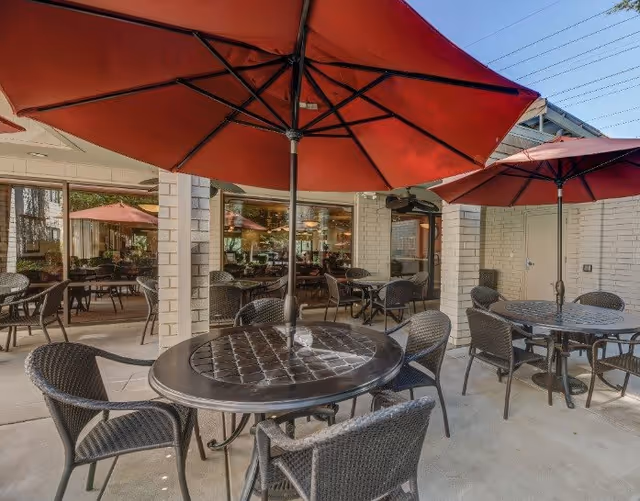 Outdoor patio with round tables, wicker chairs and red umbrellas outside a senior living building.